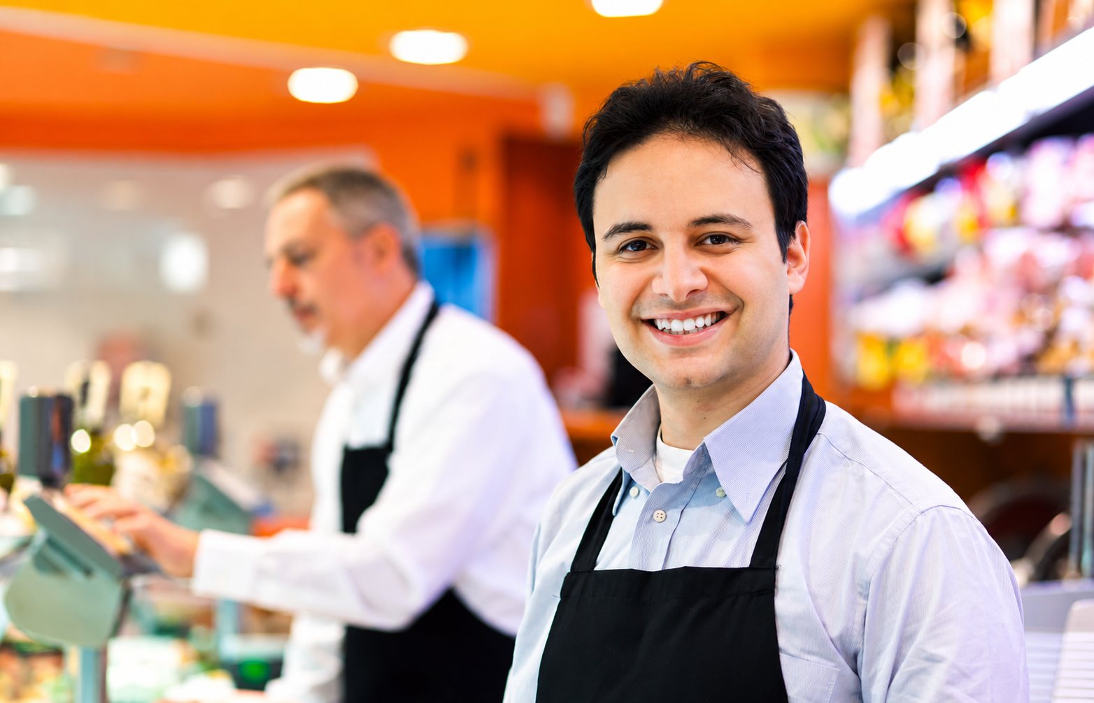 Deux commerçants en tablier dans une boutique, l’un souriant face caméra